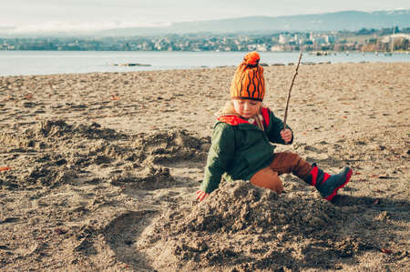 Cute toddler boy playing by the lake outside, wearing warm jacket and winter boots, happy childhoodの写真素材