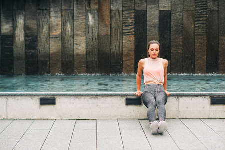 Outdoor portrait of young beautiful fit woman doing tricep dips, wearing fashionable activewear, athlete model posing in urban background, sport fashionの写真素材