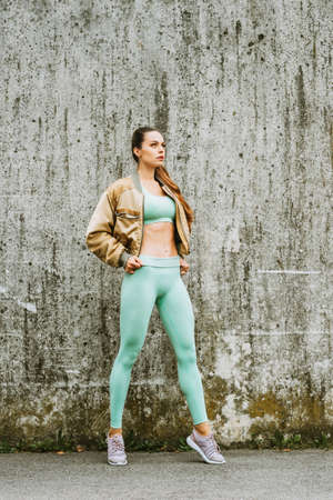 Outdoor portrait of young beautiful fit woman, wearing green activewear, athlete model posing next to grey urban wall background, sport fashionの写真素材