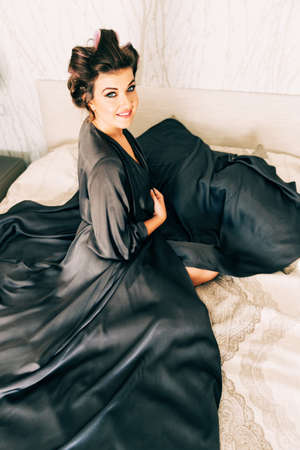 Interior portrait of happy young woman relaxing in bedroom, wearing grey nightie, curlers on the headの写真素材