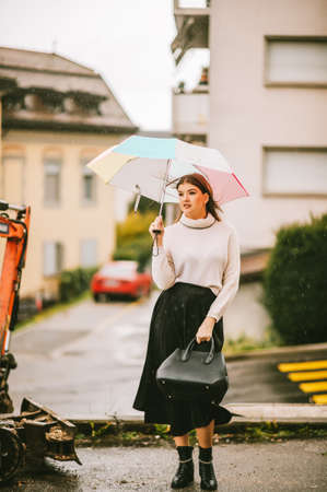 Stylish young woman under the rain, holding colorful umbrella, wearing warm pullover and black skirtの写真素材