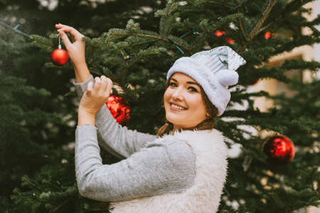 Festive portrait of happy young woman decorating Christmas tree, wearing silver Santa hatの写真素材