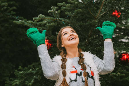 Festive portrait of happy young woman posing outside by the Christmas tree, wearing green mittensの写真素材