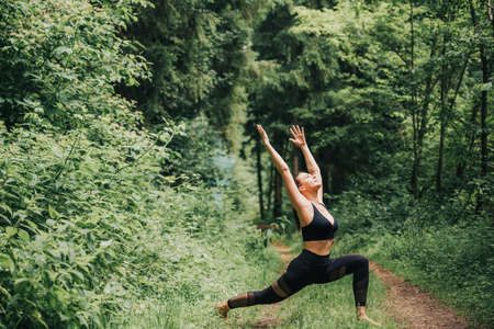 Young fit woman practicing yoga in summer forest, wearing black leggings and topの写真素材