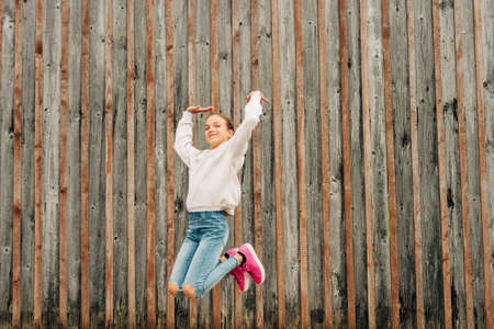 Outdoor portrait of happy young girl wearing grey sweatshirt, posing on brown background, jumping...