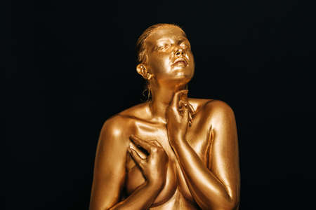 Portrait of sensual woman covered with golden paint, posing on black background, eyes closed