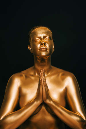 Portrait of sensual woman covered with golden paint, posing on black background, eyes closed, holding hands next to chest, namaste gesture signの写真素材
