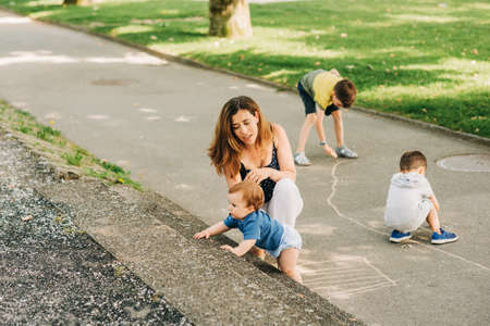 Happy mother spending time with children in summer park, playing with baby, boys drawing with chalkの写真素材