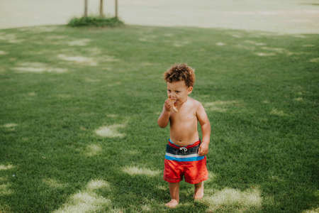 Outdoor portrait of cute african toddler boy playing by the pool, wearing swimming shorts, eating piece of pizzaの写真素材