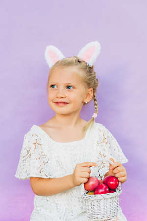 Cute little girl wearing bunny ears, holding white basket full of painted eggs, posing on pink...