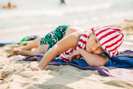 Summer portrait of cute little boy relaxing on sand beach by the sea, lying down covered with towels, vacation with childrenの写真素材
