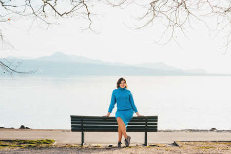 Outdoor portrait of young woman resting on the bench by the lake in early spring, wearing blue knitted pullover dressの写真素材