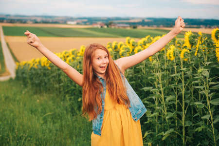 Outdoor portrait of cute young red-haired girl posing in sunflower field, holding arms up wide openの写真素材