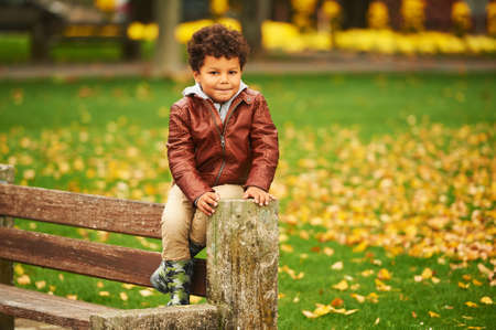 Outdoor autumn portrait of cute african boy, wearing brown leather jacket, child having fun in park, sitting on the top of the benchの写真素材