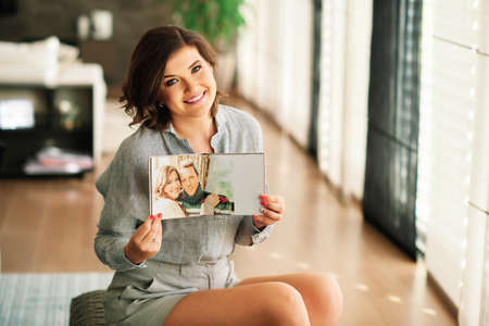 Young happy woamn looking at photo album, resting in cozy living roomの写真素材