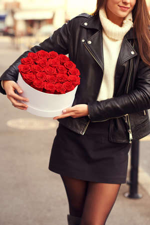 Outdoor portrait of young beautiful woman holding white box with red roses, wearing leather jacketの写真素材