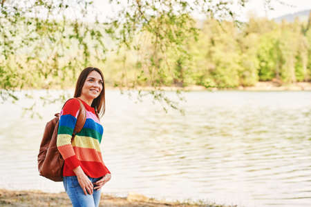 Outdoor portrait of young beautiful woman relaxing by river on a nice warm day, wearing colorful...