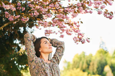Outdoor spring portrait of pretty young woman with brown hair posing under pink blossoming treeの写真素材