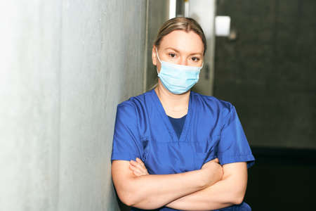 Young female scrub nurse wear blue uniform and face mask, standing in hospital hallway, leaning on grey wallの写真素材