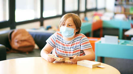 Little boy sitting in a classroom or library, wearing face mask, holding a book, back to school...