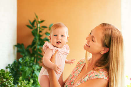 Happy young mother playing with adorable baby girl, family at home, cozy green balcony with many...