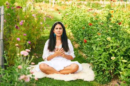 Middle age woman meditating in green garden, holding hands on heart, eyes openの写真素材