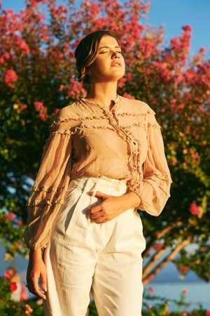 Outdoor fashion portrait of stylish young woman wearing beige blouse and white trousers, posing...