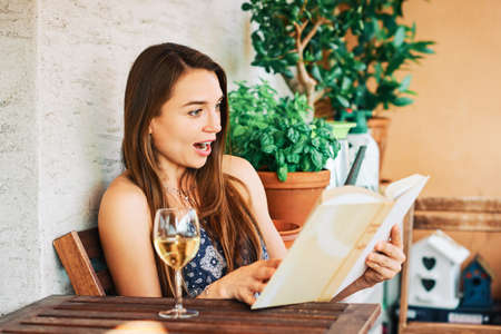 Young happy woman relaxing on the balcony, reading interesting book, drinking glass of wineの写真素材