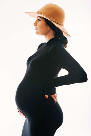 Studio maternity portrait of gorgeous young woman, posing on white background, wearing black turtle neck dress and a hatの写真素材