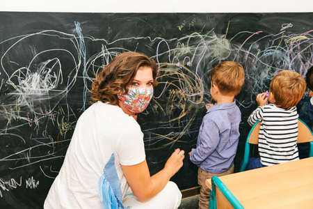 Art classroom in kindergarten, young teacher drawing with children on chalk wall, wearing facemaskの写真素材