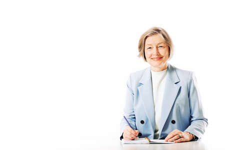 Studio portrait of middle age woman posing on white background, sitting at the desk and taking notes