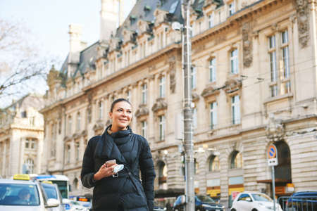 Happy young woman walking down the street, wearing warm black jacket, holding phone and facial mask in handsの写真素材
