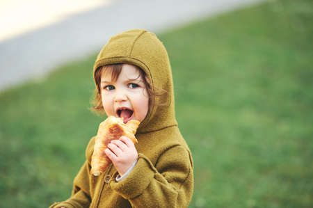 Outdoor portrait of cute 1 - 2 year old toddler kid eating croissant