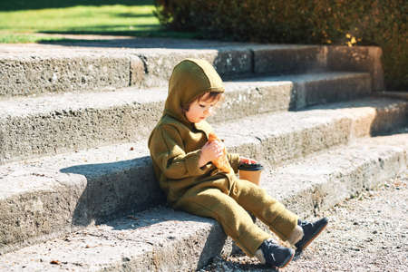 Funny little kid eating croissant and drinking from takeaway coffe cup in early morning, sitting on...