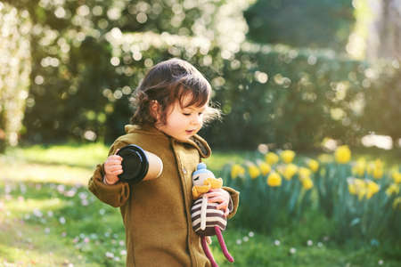 Outdoor portrait of adorable toddler girl playing in spring park, holding cup of takeaway coffee and...