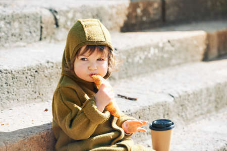 Funny little kid eating croissant and drinking from takeaway coffee cup in early morning, sitting on stairs in public parkの写真素材