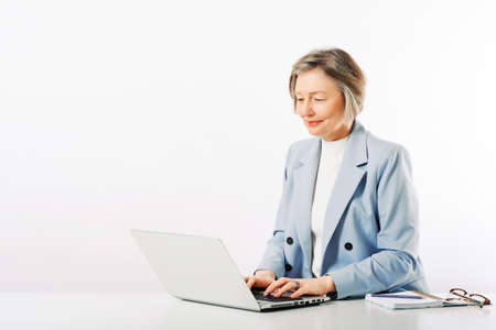 Studio portrait of middle age woman posing on white background, sitting at the desk, using laptopの写真素材