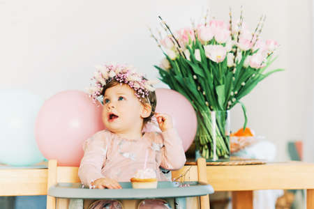 Adorable baby girl sitting in a chair, looking at cupcake with one candle, first birthday partyの写真素材