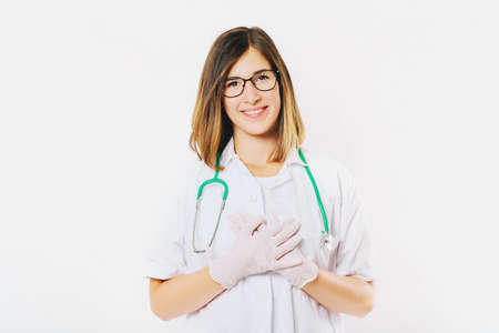Studio portrait of young female doctor posing on white background, holding hands on chest next to heart, thank you signの写真素材