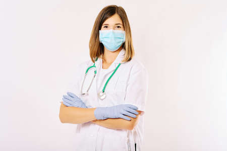Studio portrait of young female doctor posing on white background, wearing facial mask and gloves, arms crossedの写真素材