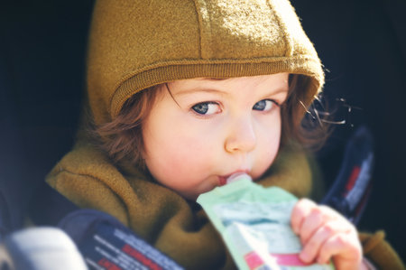 Close up portrait of sweet toddler kid eating fruit puree from plastic doy pack, sitting in stroller, outdoor snack timeの写真素材