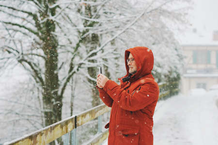 Middle age man holding smartphone, taking pictures or video for social media, winter weather with snowfallの写真素材