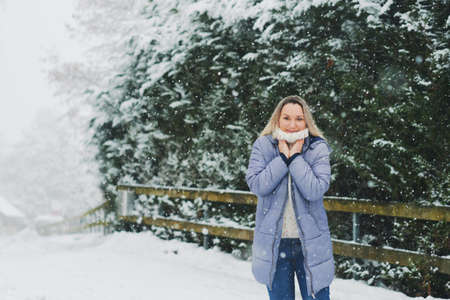 Happy mature woman enjoying nice winter day, wearing warm white pullover and blue winter jacket, standing under snowfallの写真素材