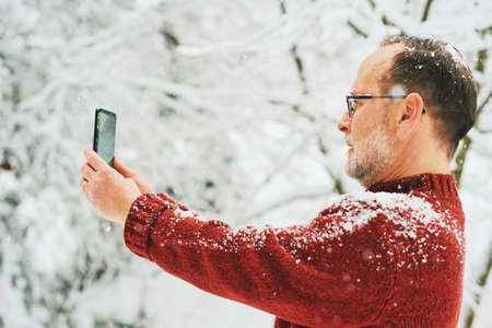 Middle age man holding smartphone, taking pictures or video for social media, winter forest with snowfallの写真素材