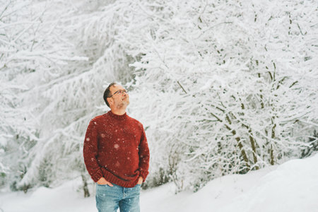 Outdoor portrait of middle age 55 - 60 year old man enjoying nice cold weather in winter forest, wearing red knitted pulloverの写真素材