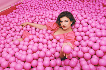 Fashion portrait of happy young woman playing in a ball pit, wearing pink dressの写真素材