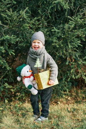 Outdoor portrait of happy sweet little boy posing next to pine tree, holding gift box and snowman toy, wearing worm scarf and hatの写真素材