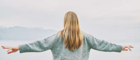 Panoramic banner background back view of young blond woman with arms wide open enjoy landscape of cloudy mountainsの写真素材