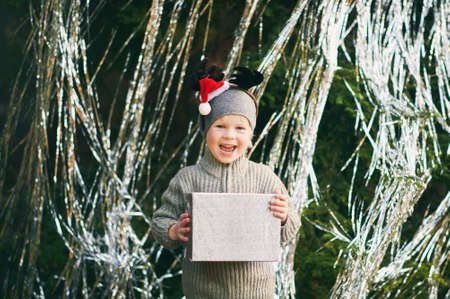 Outdoor portrait of happy and excited little boy posing next to decorated pine tree, holding gift...