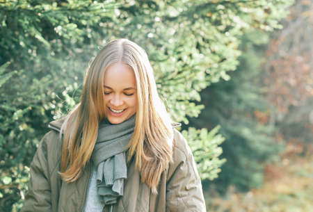Outdoor portrait of beautiful blond woman, lokking down, wearing warm scarf and winter jacket, walking in pine forestの写真素材
